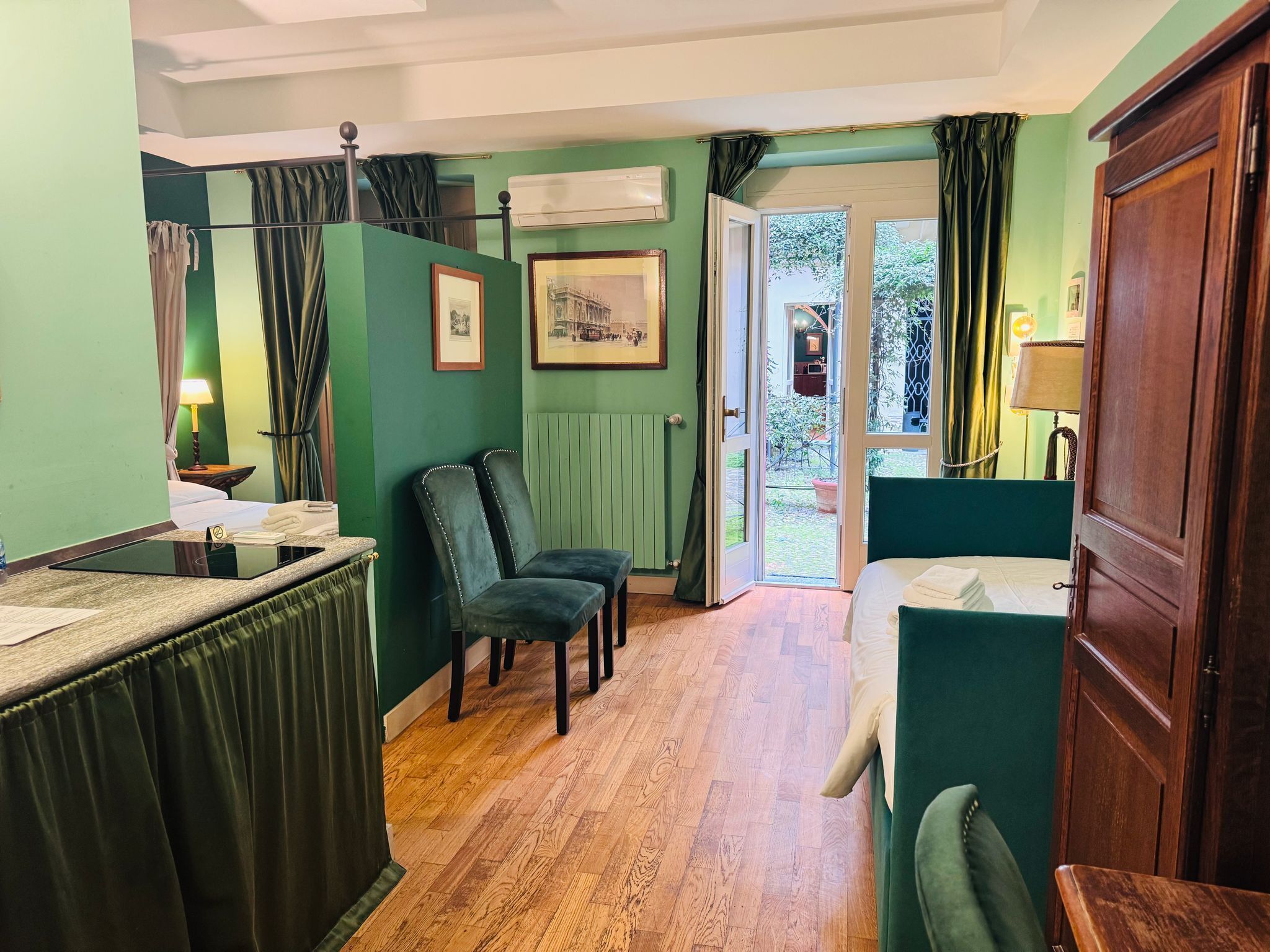 A room with a bed, a desk, two chairs, a radiator, and a door leading to a garden.