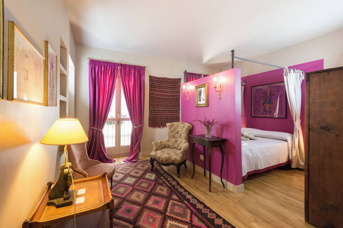 Un dormitorio con una cama, una silla, una mesa, una lámpara y una alfombra estampada.