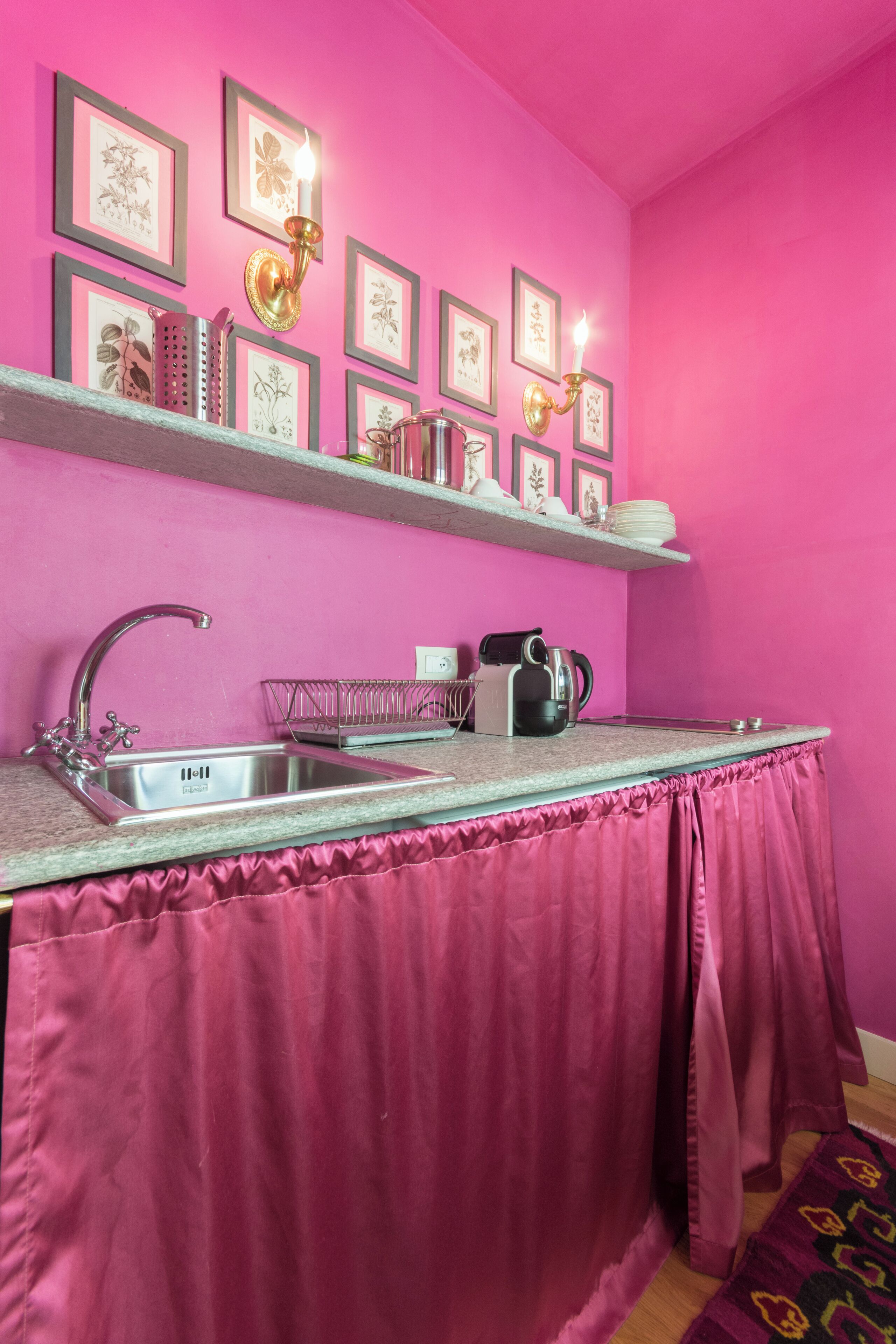 A kitchen with a pink wall, a stainless steel sink, a kettle, and framed botanical prints on shelves.