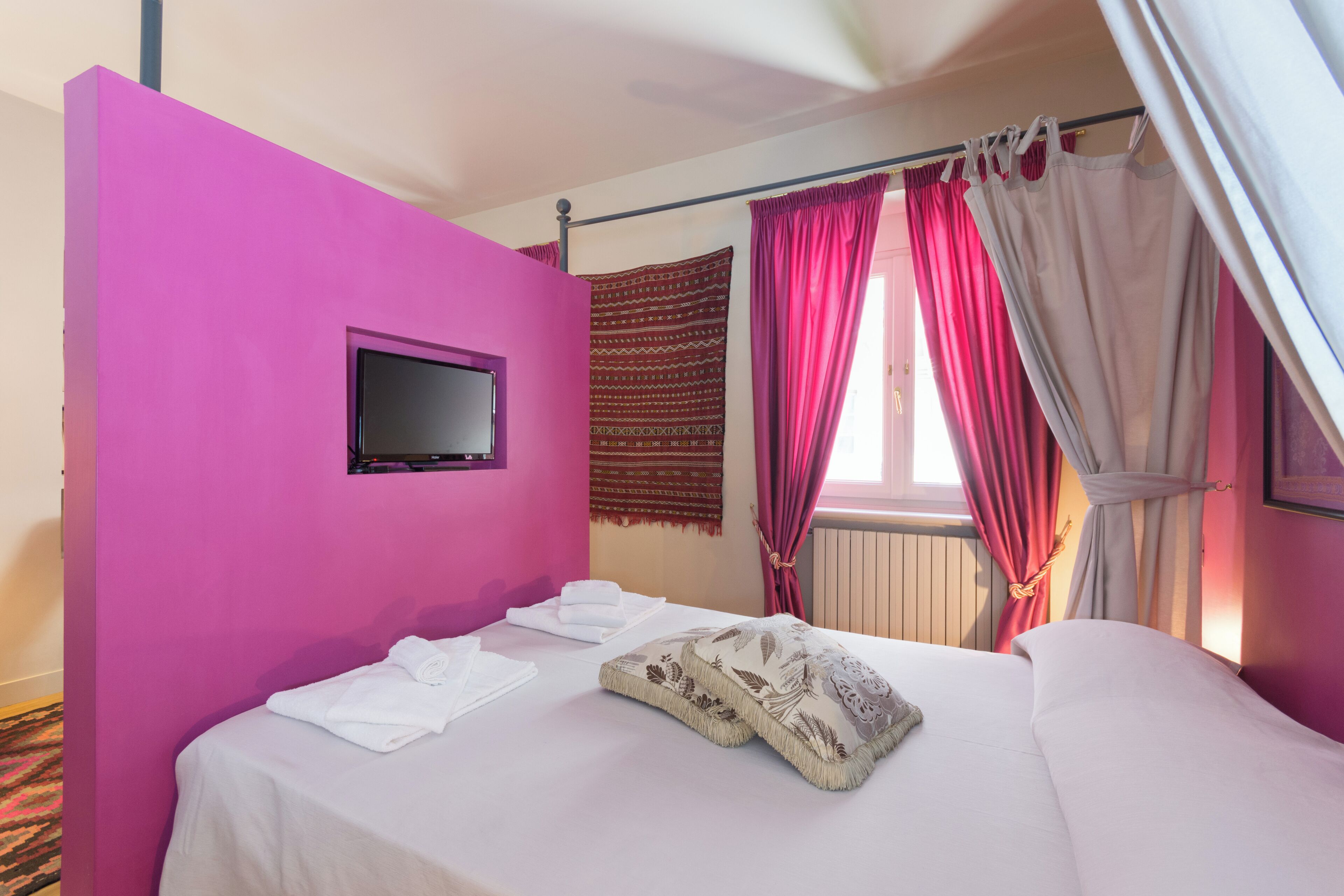 Una habitación de hotel con pared rosa, una cama con ropa blanca, un televisor de pantalla plana montado en la pared y una ventana con cortinas rojas.