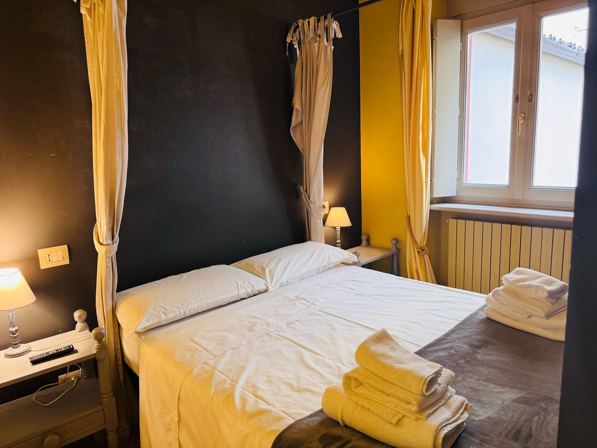 Una cama bien tendida con sábanas blancas, una mesita de noche con una lámpara y una ventana con cortinas.