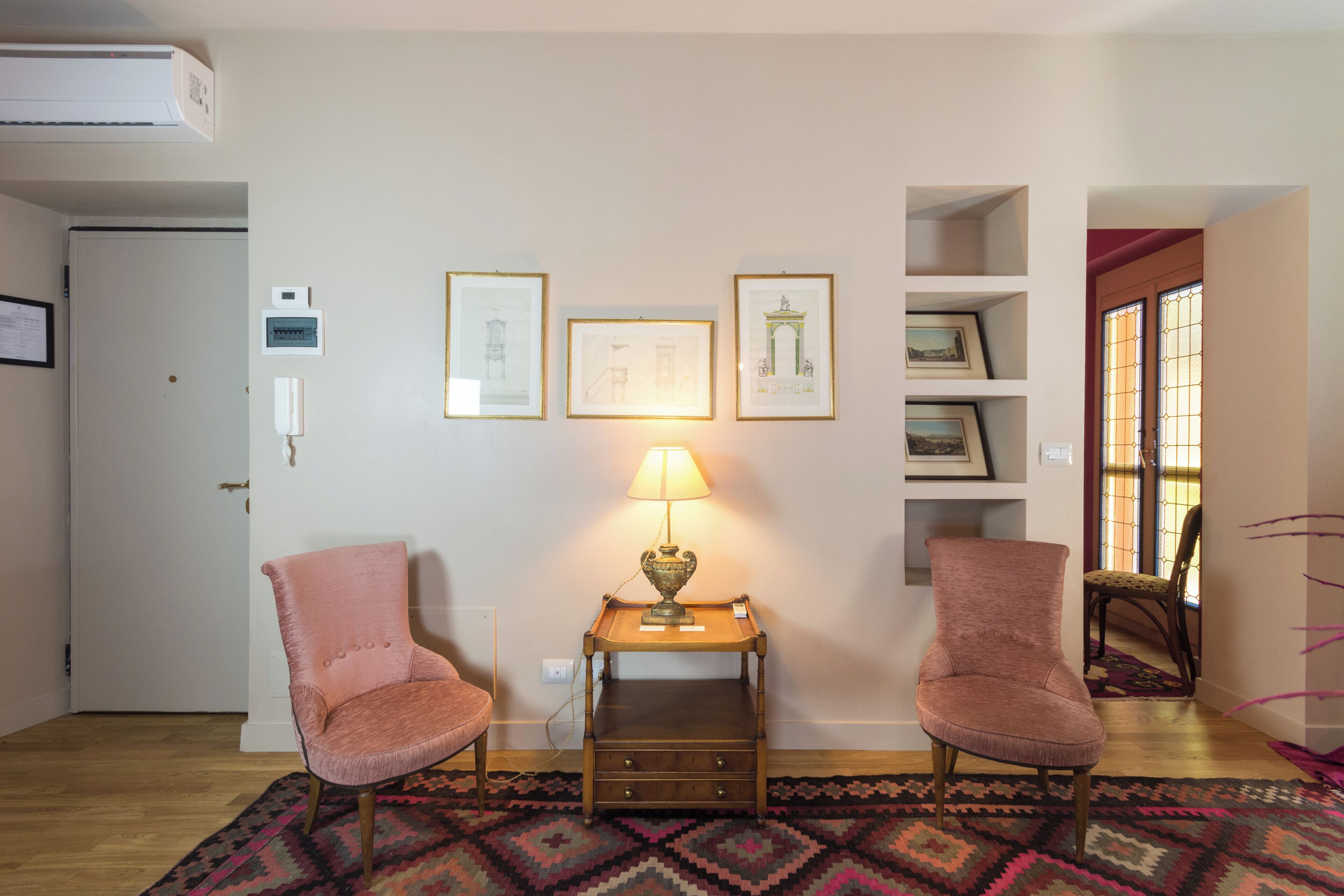 Una habitación con dos sillas rosadas, una mesita con una lámpara, cuadros en la pared y una alfombra estampada.