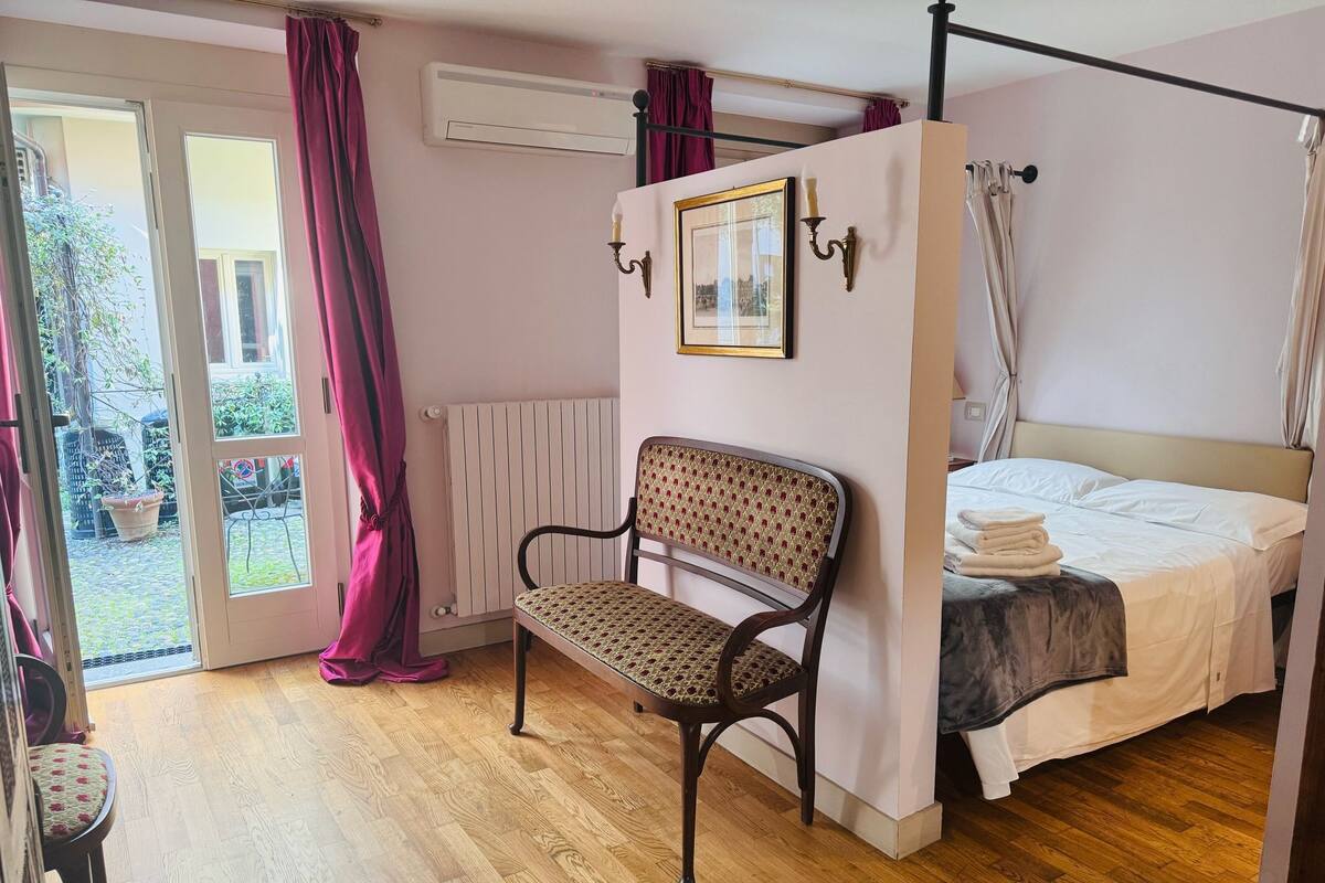 Un dormitorio con una cama con dosel, una silla, un radiador y una ventana con cortinas.