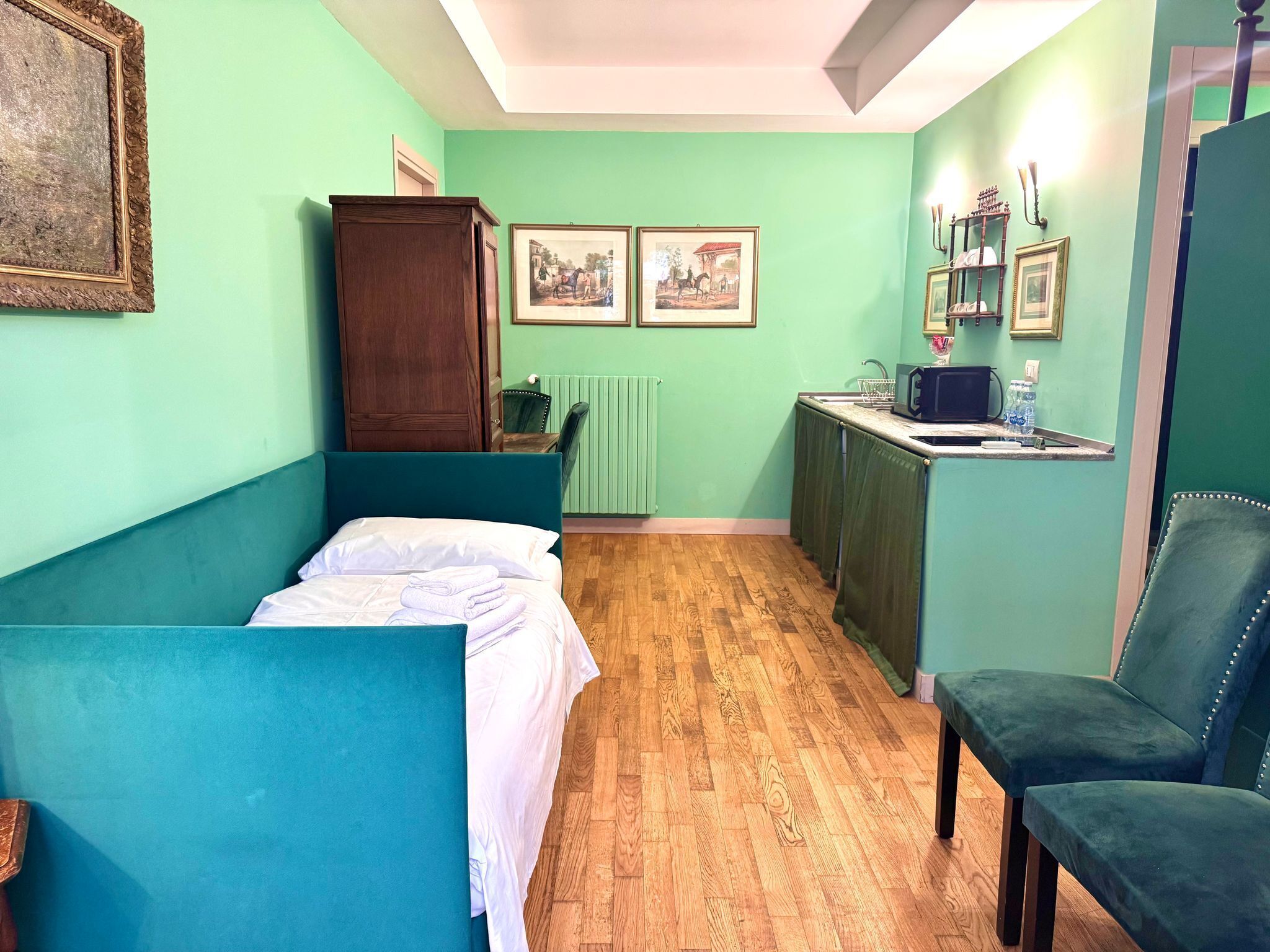 A small, cozy room with a bed, a chair, a wooden cabinet, and a kitchenette.