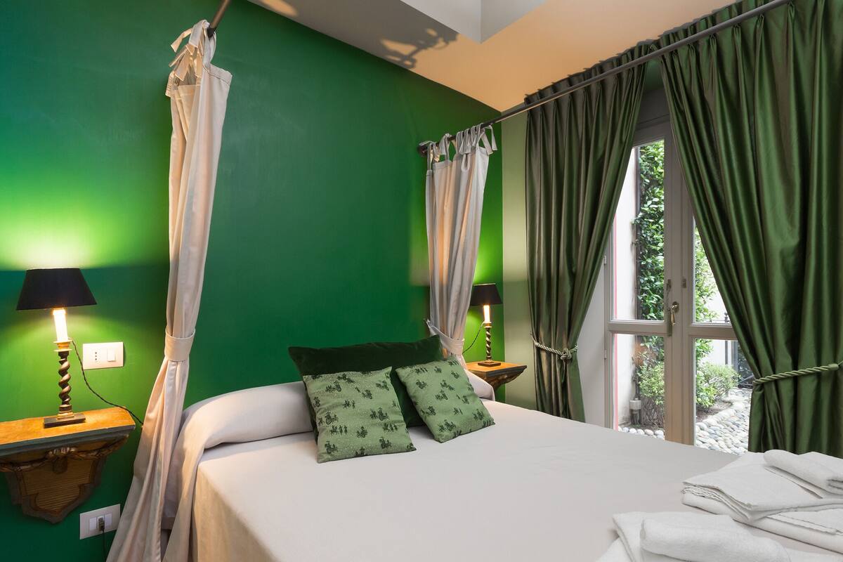Un dormitorio con pared verde, una cama con ropa de cama blanca, dos almohadas estampadas en verde y una ventana con cortinas verdes, además de dos lámparas de noche.