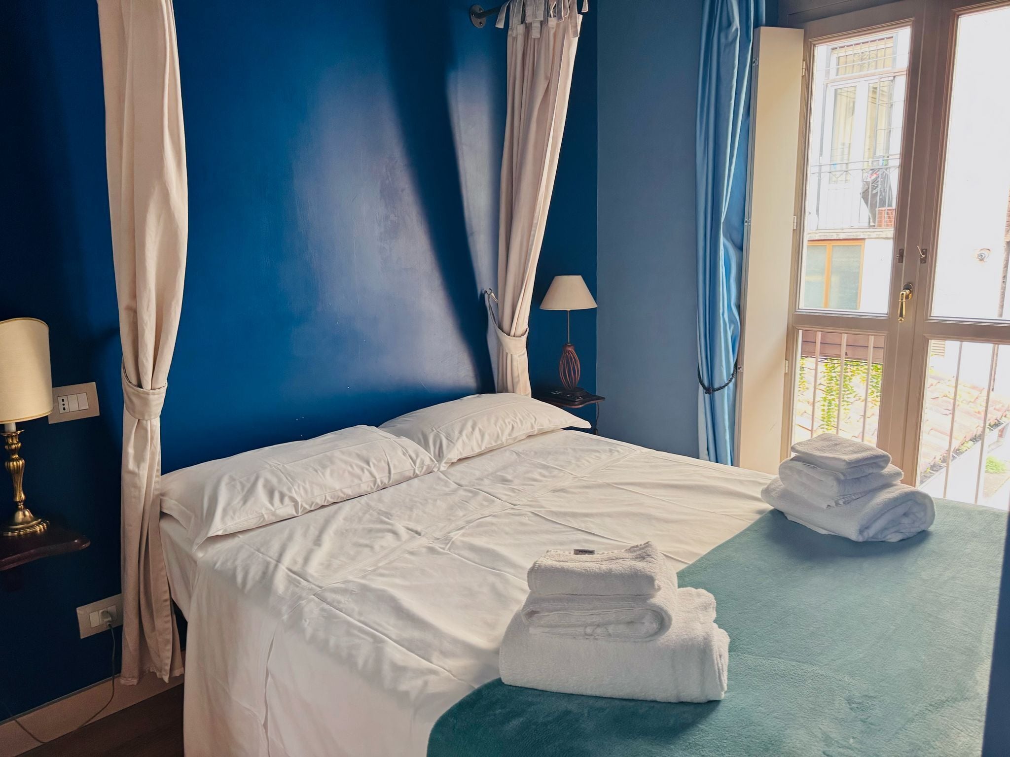 A bedroom with a bed, curtains, a lamp, and towels on a green mat.