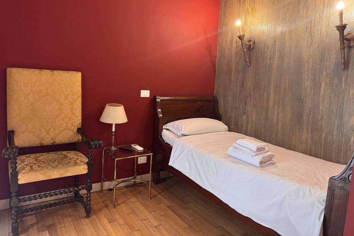 Una cama individual con ropa de cama blanca, una mesita de noche de madera con lámpara, una silla con cojín estampado y una pared roja con sección texturizada.