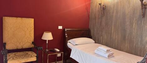 A single bed with white linens, a wooden nightstand with a lamp, a chair with a patterned cushion, and a red wall with a textured section.
