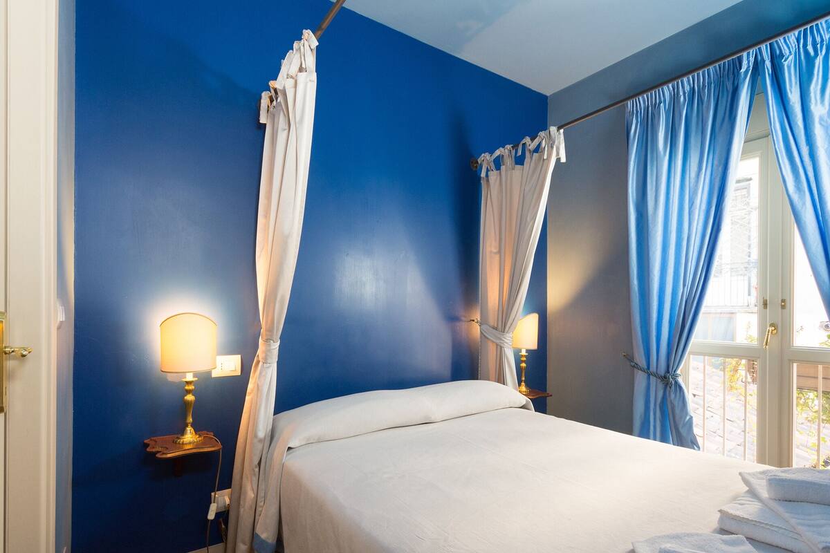 Un dormitorio con una cama, dos lámparas de noche, un ventanal con cortinas y una pila de toallas.