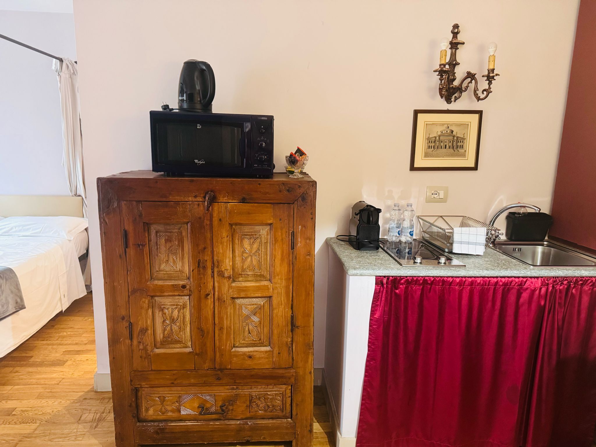 A compact hotel room with a wooden cabinet, a microwave, a sink with a red curtain, a chandelier, and a framed picture on the wall.
