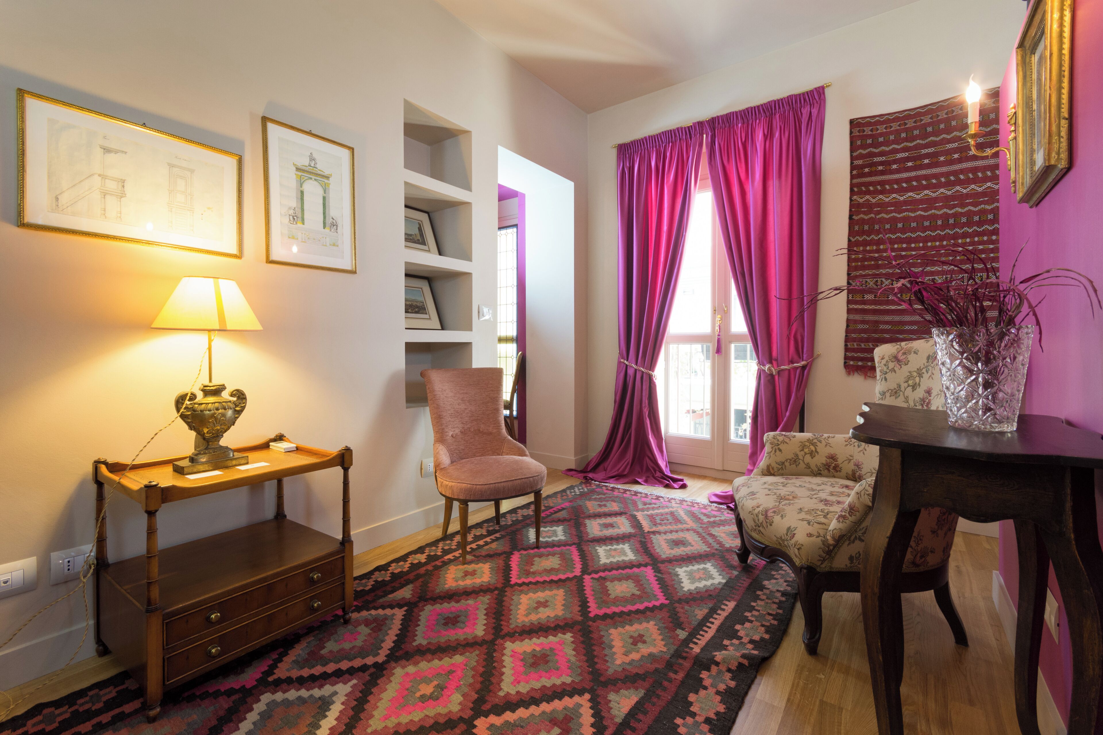 A room with a wooden side table, a lamp, a patterned rug, a chair, a bookshelf, and a window with purple curtains.