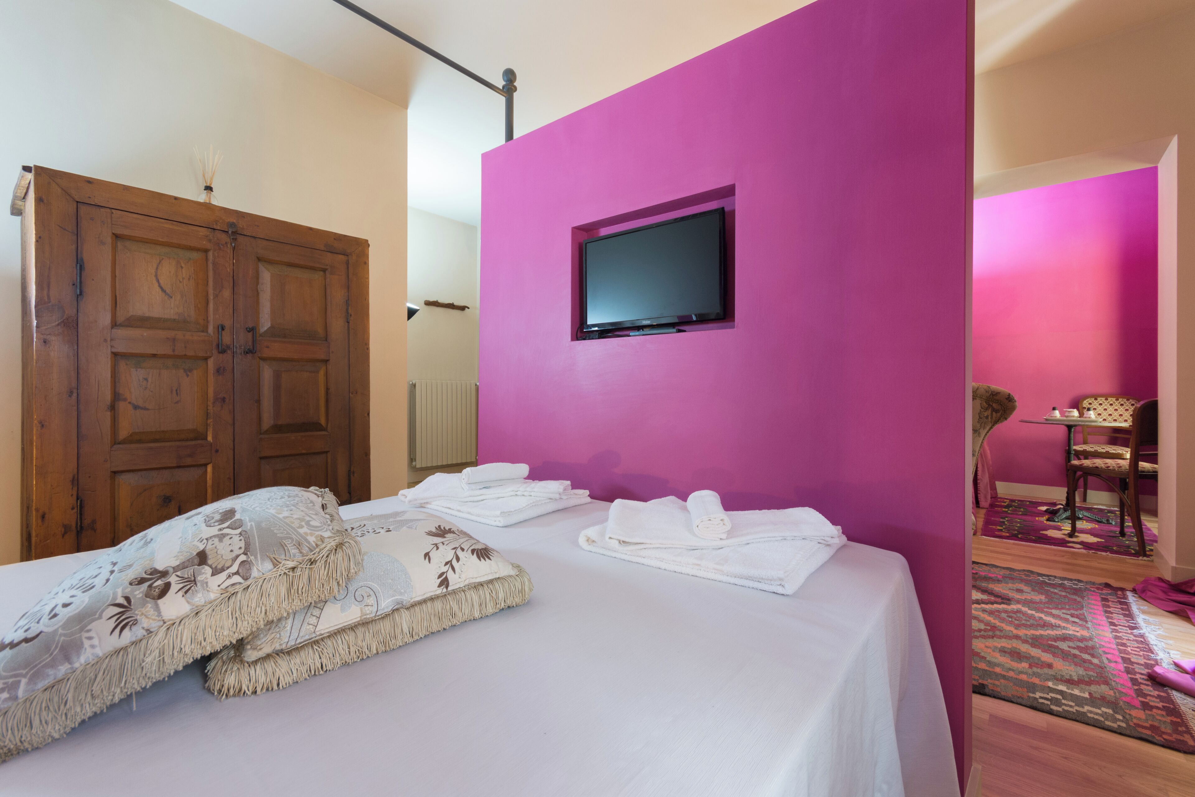 A bedroom with a bed, a television mounted on a pink wall, a wooden wardrobe, and a chair in the corner.