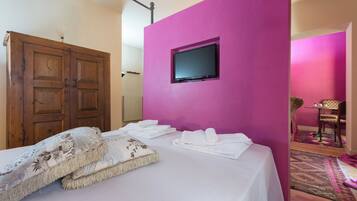 A bedroom with a bed, a television mounted on a pink wall, a wooden wardrobe, and a chair in the corner.