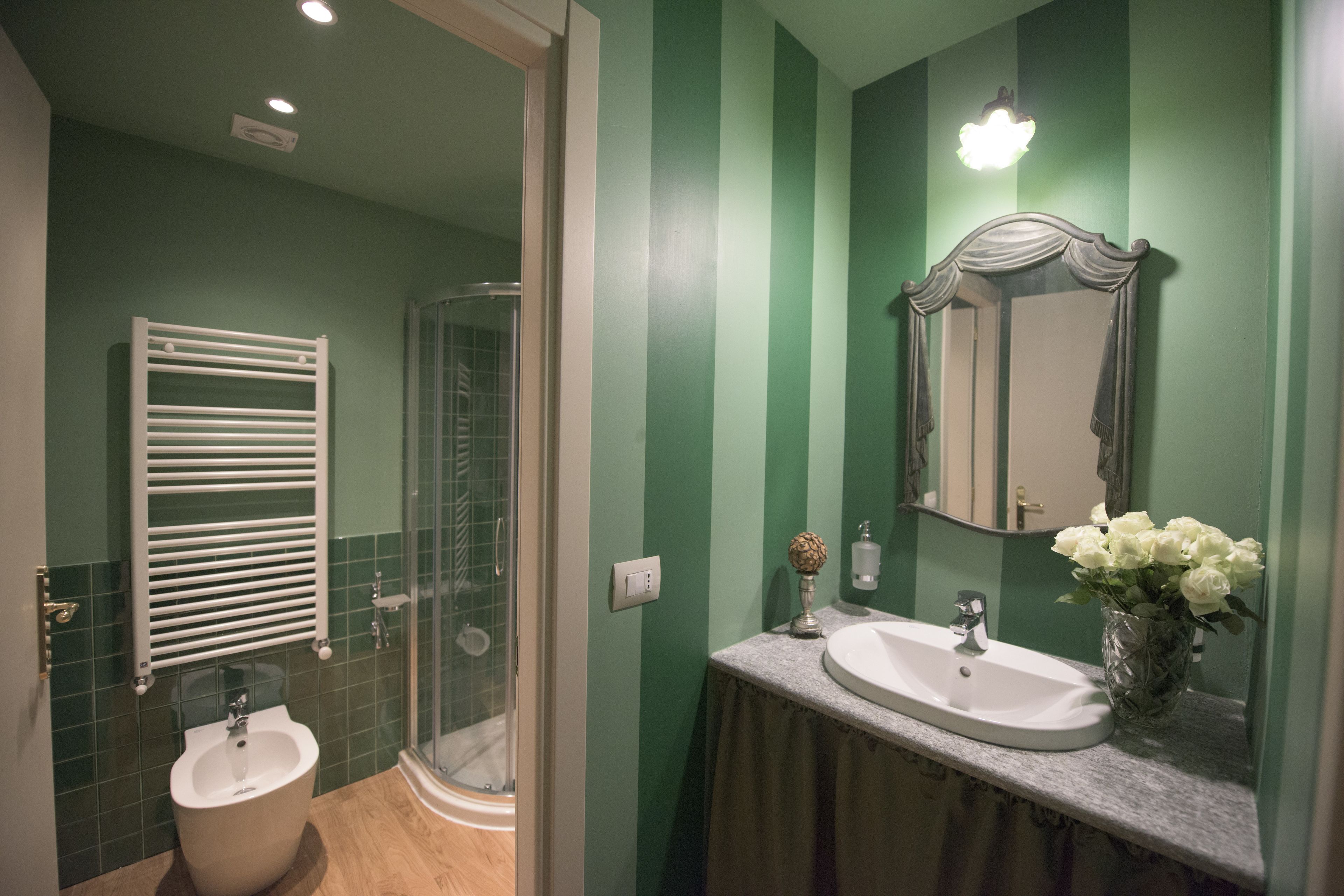 A bathroom with a glass shower enclosure, a white pedestal sink, a vanity with a mirror, and a vase with flowers.