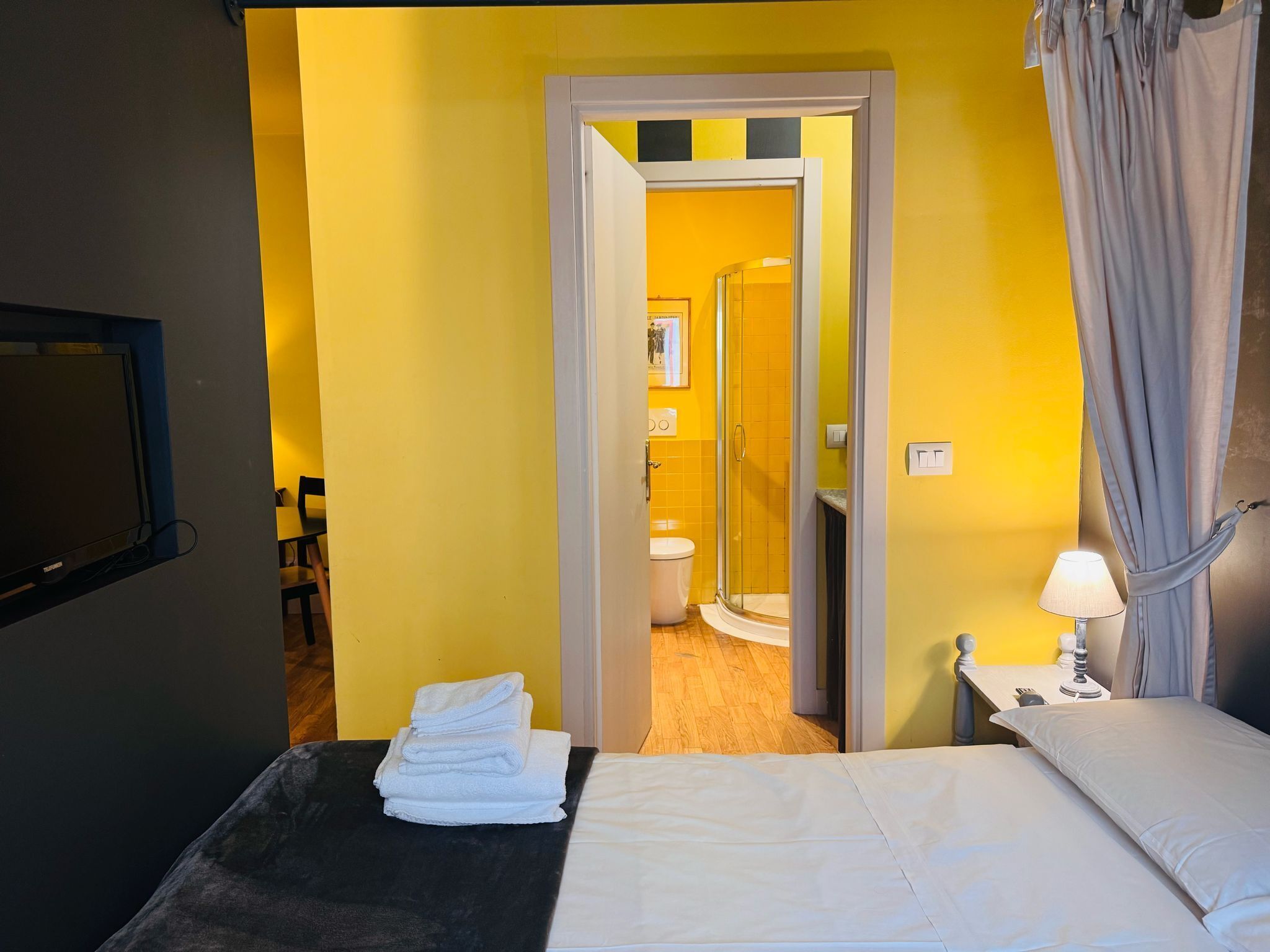 A hotel room with a bed, a small table with a lamp, a TV, and a bathroom visible through an open door.