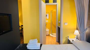 A hotel room with a bed, a small table with a lamp, a TV, and a bathroom visible through an open door.