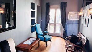 A room with a blue armchair, a wooden table with papers, a sofa, a wicker chair, and a large window with curtains. - Residence Porta Palace - Welkome Apartments (Turin)