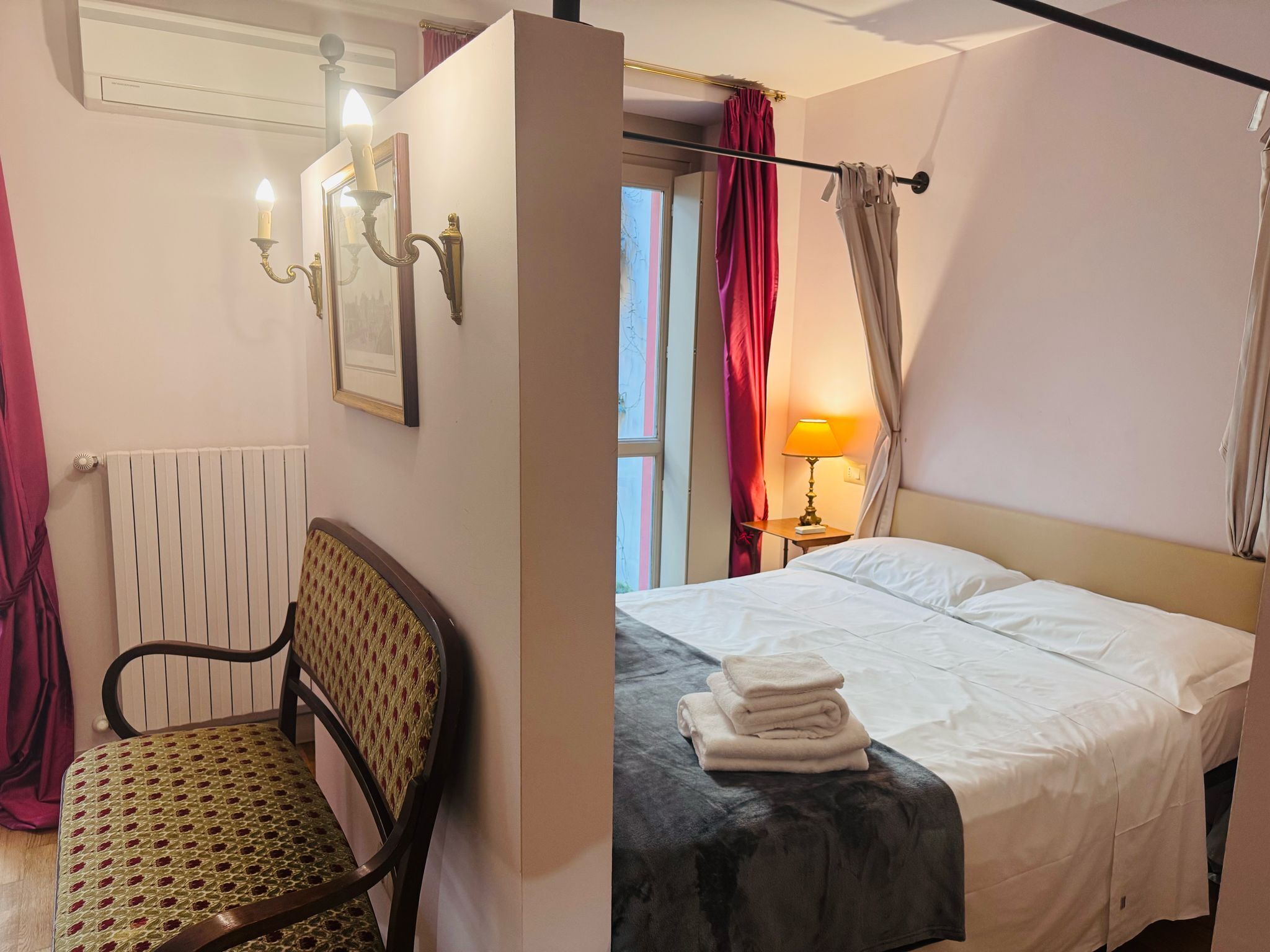 Una habitación de hotel con cama, una silla, un radiador, una lámpara y cortinas.