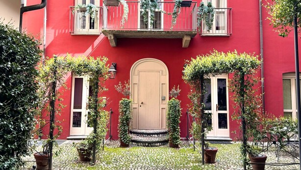 A red building with multiple windows and balconies, some with plants. There are potted plants in front of the building and a cobblestone courtyard. - Residence Porta Palace - Welkome Apartments (Turin)