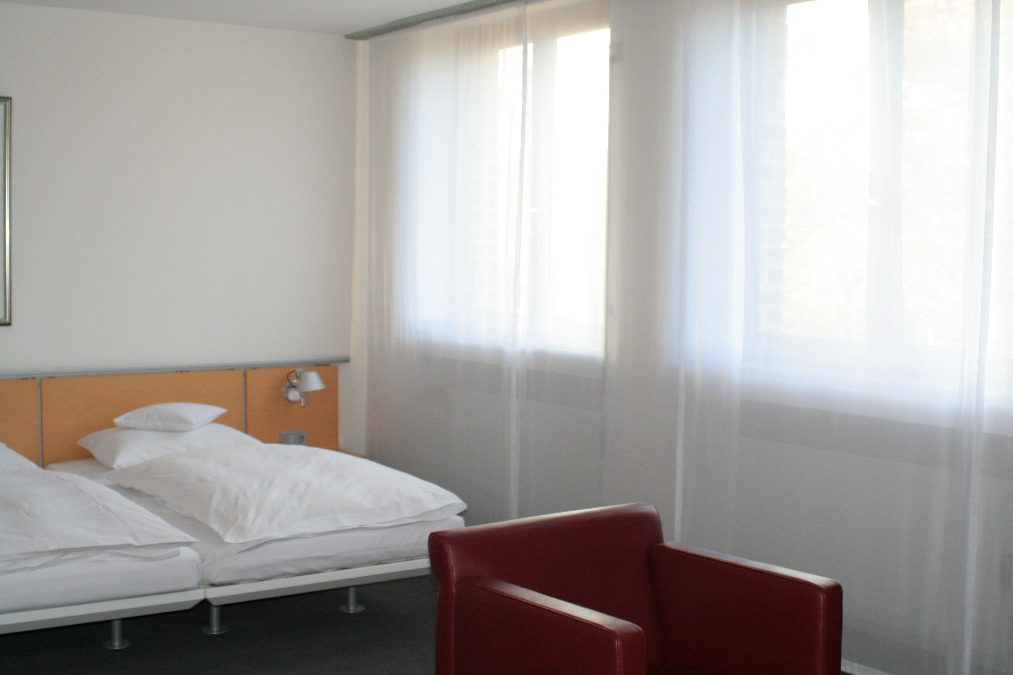 A hotel room with two beds, a red chair, and a window with sheer curtains.