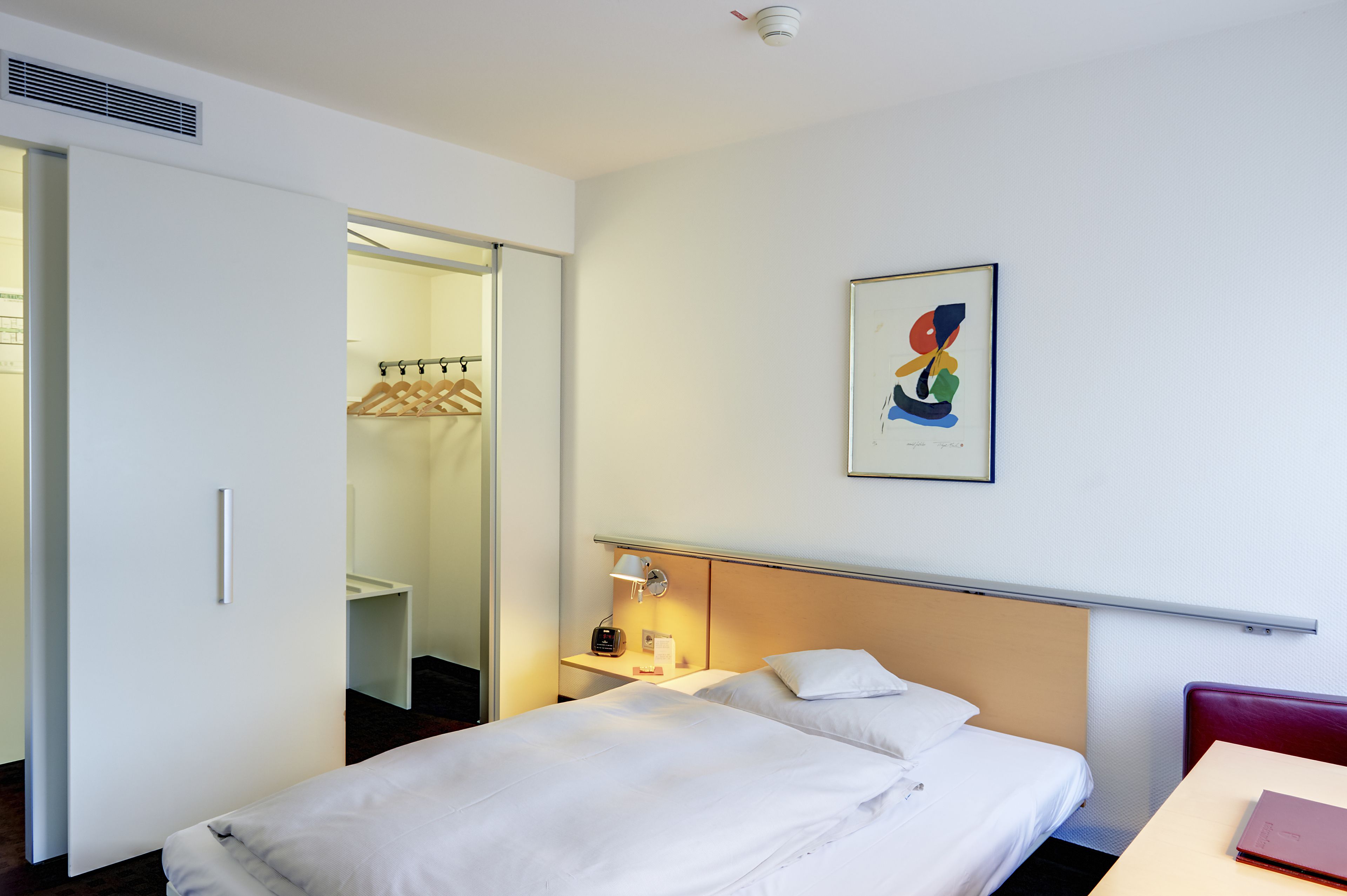 Een hotelkamer met een bed, nachtkastje, lamp, rode stoel, garderobe en een schilderij aan de muur.