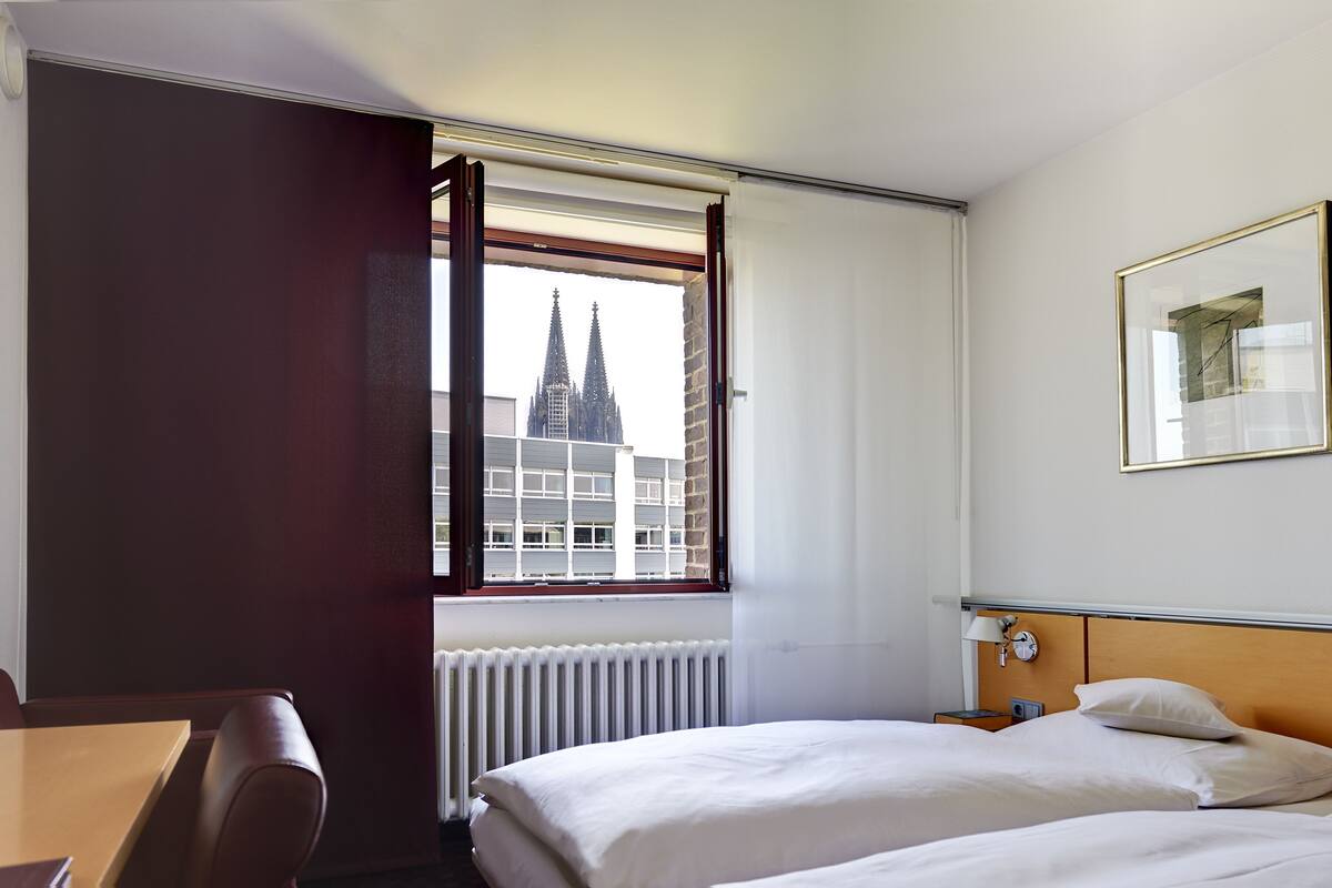 A hotel room with a bed, a desk, a chair, a window with a view of a building, and a framed picture on the wall.