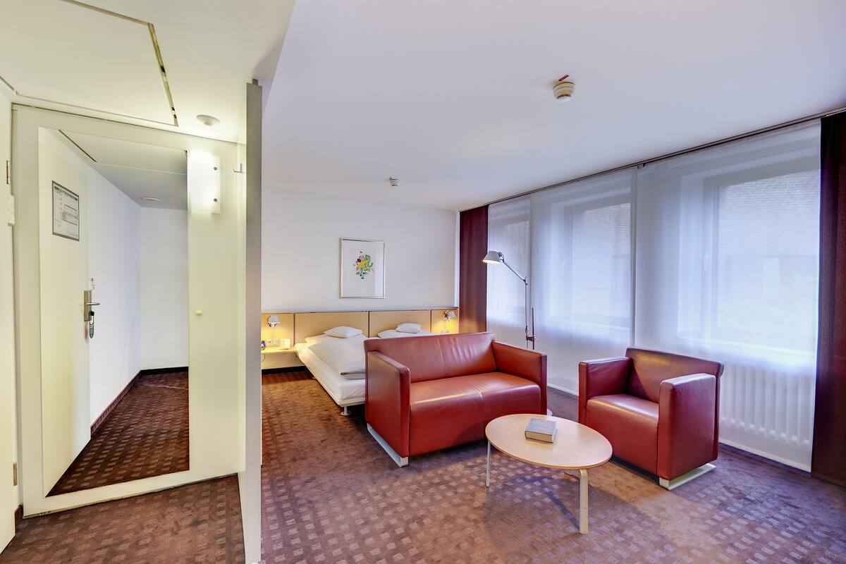 A hotel room with a bed, a red leather sofa, a small round table, and large windows with curtains.