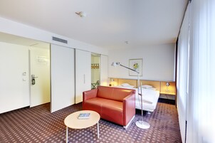 Habitación de hotel con un sofá de cuero rojo, una mesita redonda pequeña, una cama con ropa blanca, una lámpara de noche y un cuadro en la pared.