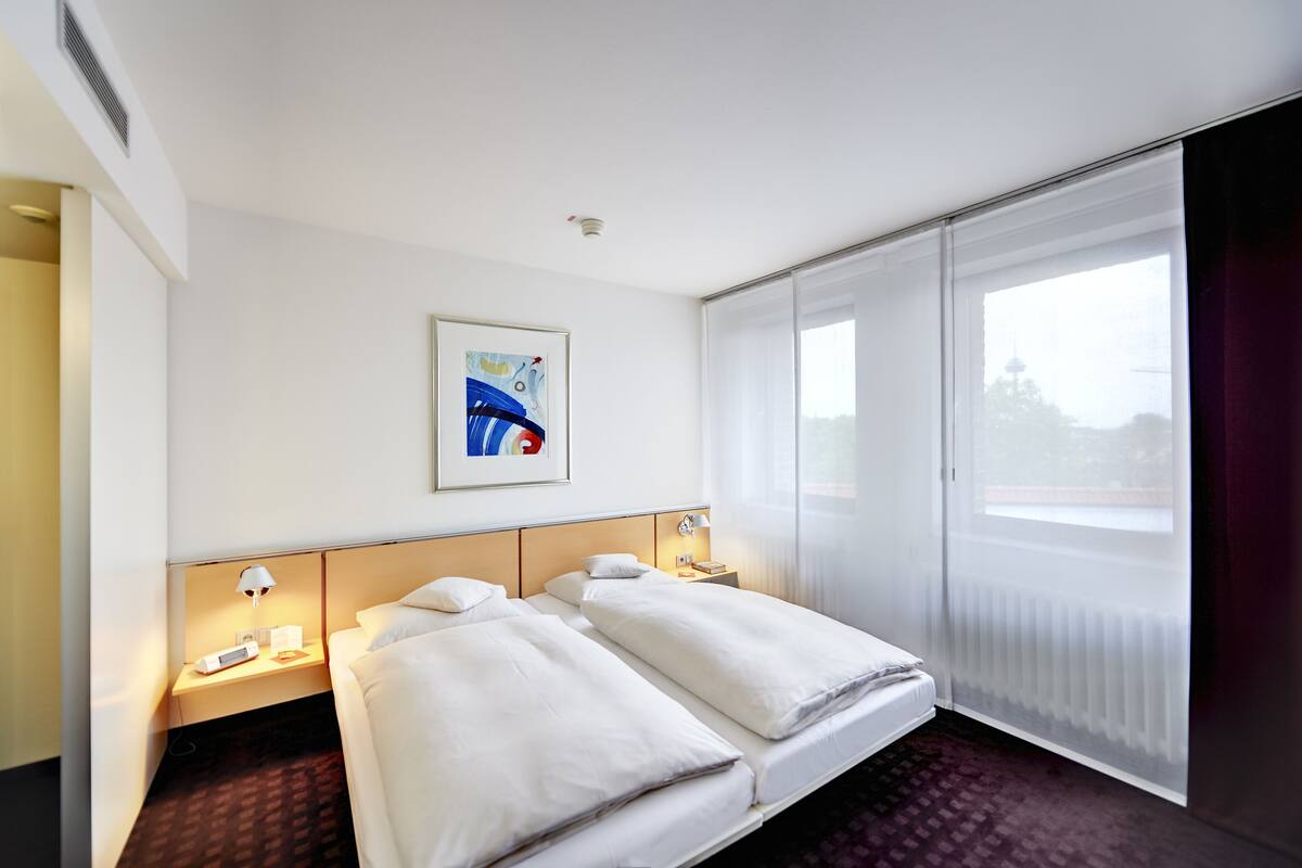 A hotel room with two beds, a large window with blinds, a painting on the wall, and a nightstand with a lamp.