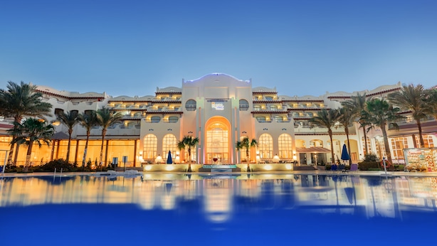 Ein Hotel mit großem, spiegelnden Pool, Palmen und einer gut beleuchteten Fassade.