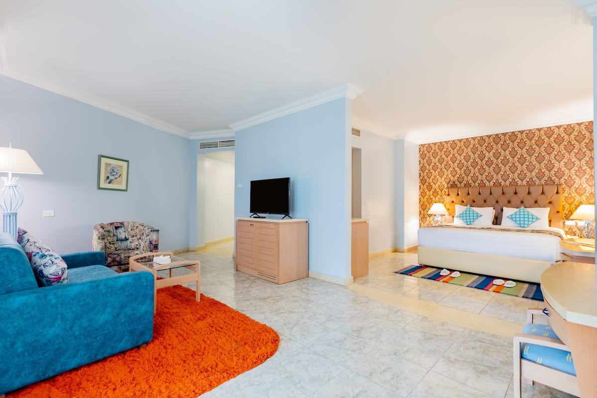 Una habitación de hotel con un sofá azul, un aparador de madera, un televisor de pantalla plana, una cama con papel tapiz estampado y una pequeña zona de comedor.