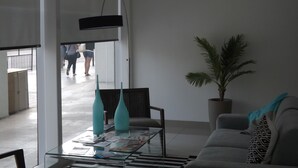 A modern living room with a glass coffee table, a sofa, a potted plant, and a large window overlooking a public area. - Edificio Vive Viana I (Vina del Mar)