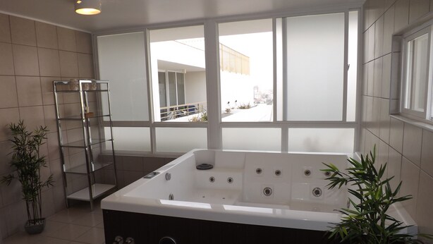 A modern bathroom with a large bathtub, a glass shelf unit, and two potted plants. - Edificio Vive Viana I (Vina del Mar)