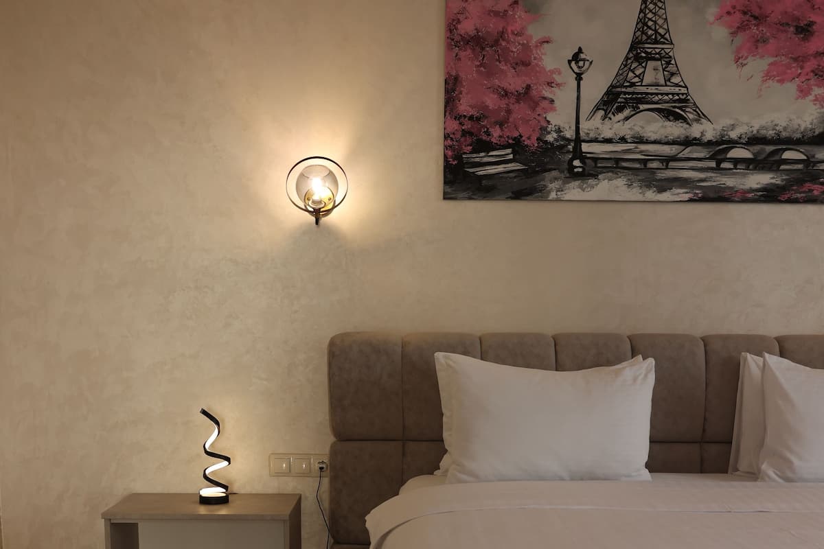 A bedroom with a large bed, a nightstand with a lamp, and a painting of the Eiffel Tower.
