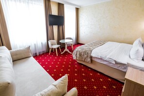 A hotel room with a red carpet, a bed, a sofa, a TV, and a small table with chairs.