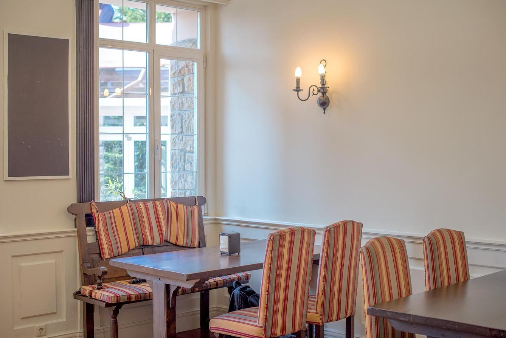 Un comedor con una mesa de madera y sillas tapizadas a rayas, una lámpara fijada a la pared y un ventanal con vistas a edificios.