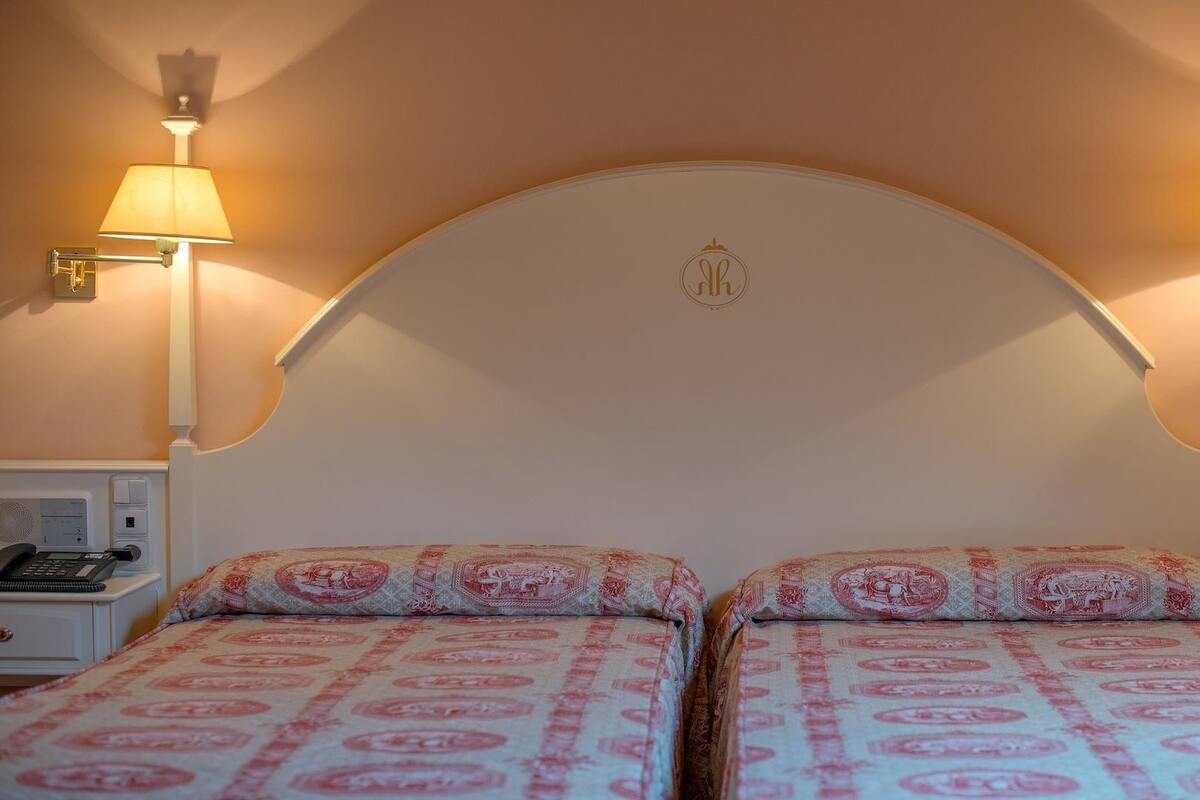 A hotel room with two beds, each with a patterned bedspread and matching pillows. There are two bedside tables with lamps and a telephone on one of them. The room has a curved headboard with a logo in the center.