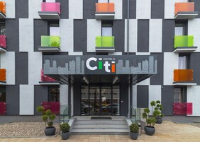 Ingresso moderno di un hotel con insegna che mostra il panorama cittadino, diversi balconi con ringhiere colorate e piante in vaso all'ingresso.