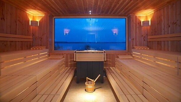 A sauna with wooden walls and benches, a large window, and two wall-mounted lamps. - See- und Waldresort Gröbern (Muldestausee)