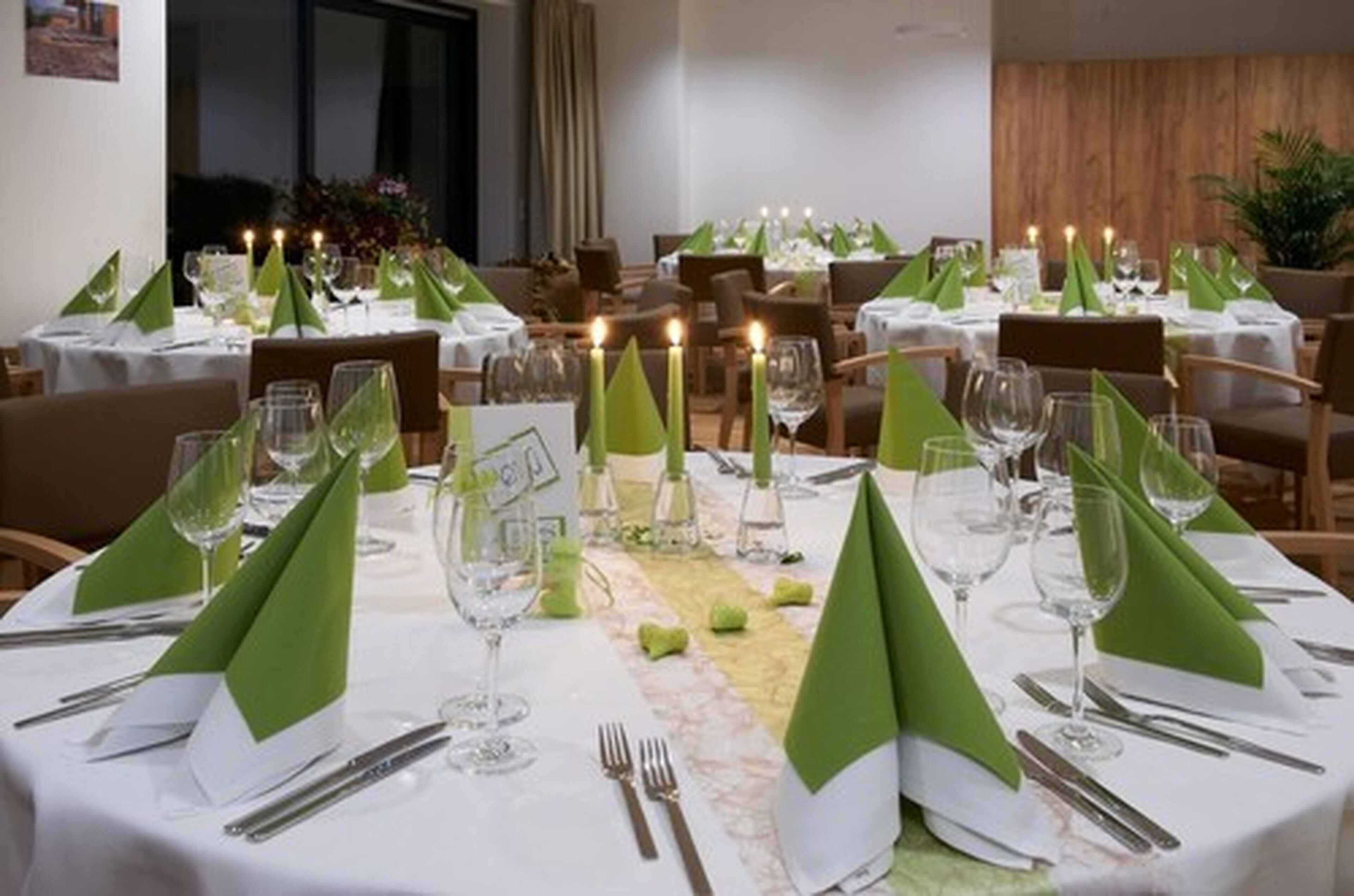 A dining area with round tables set for a meal, each with green napkins, wine glasses, and cutlery.