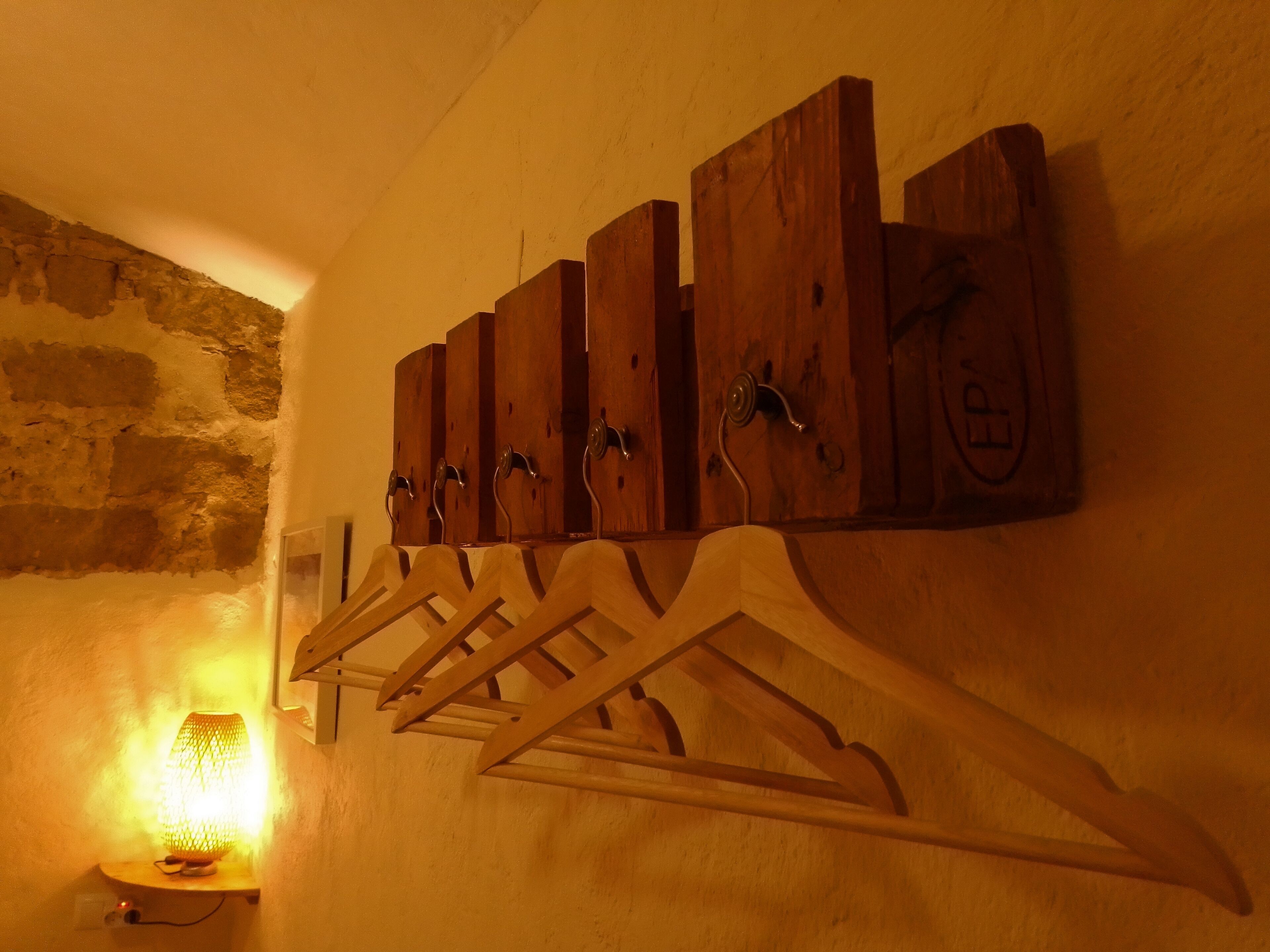 Un perchero de madera con ganchos frente a una pared, con una pared de piedra al fondo y una lámpara sobre una mesita.