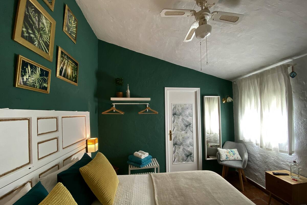 Ein Schlafzimmer mit Bett, Deckenventilator, Nachttisch, Stuhl, einem Fenster mit Vorhängen und einer Tür mit spiegelnder Blattmuster-Verzierung.