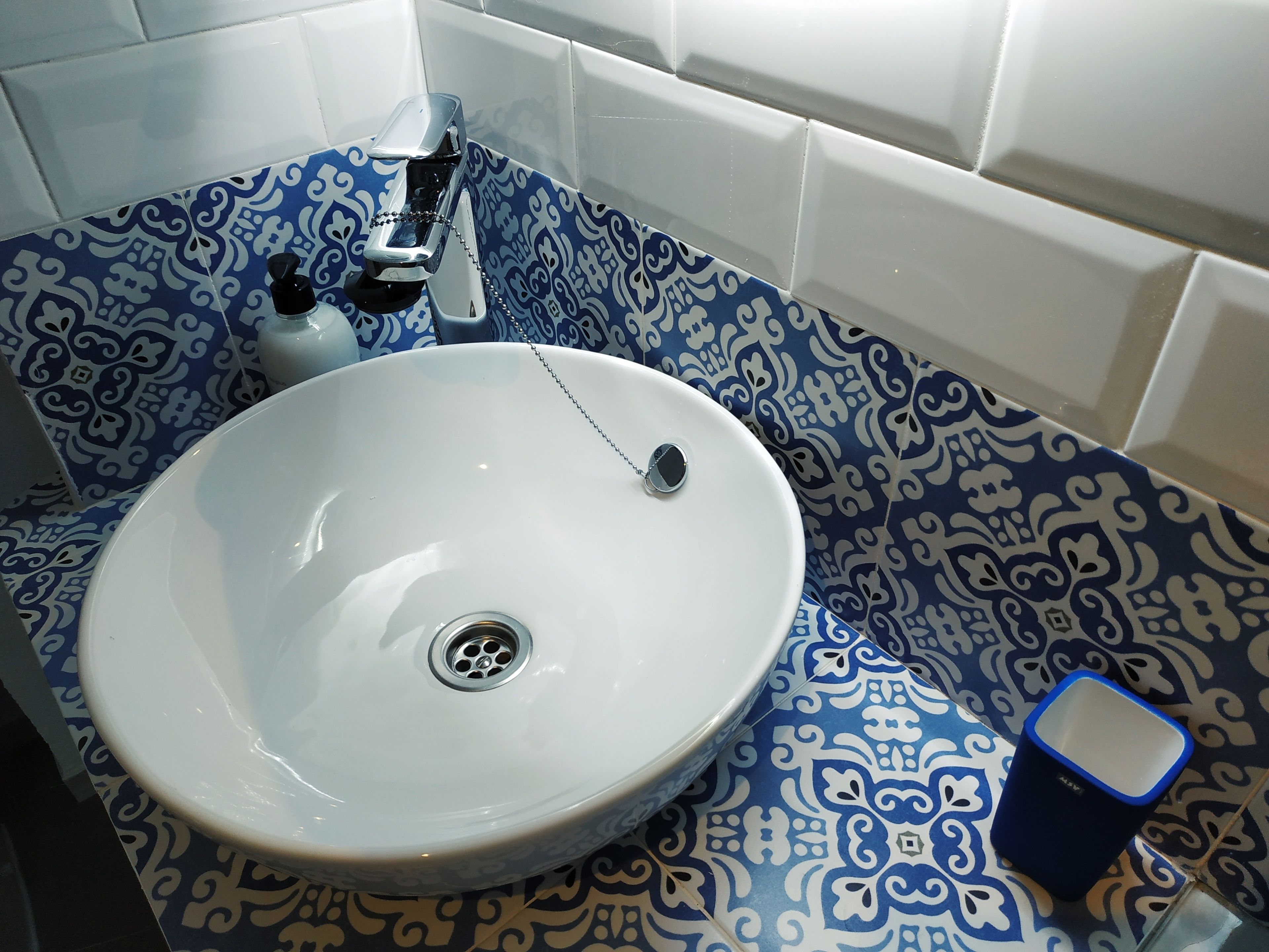 Un baño con un lavamanos blanco, baldosas azules estampadas y un cubo de basura azul.