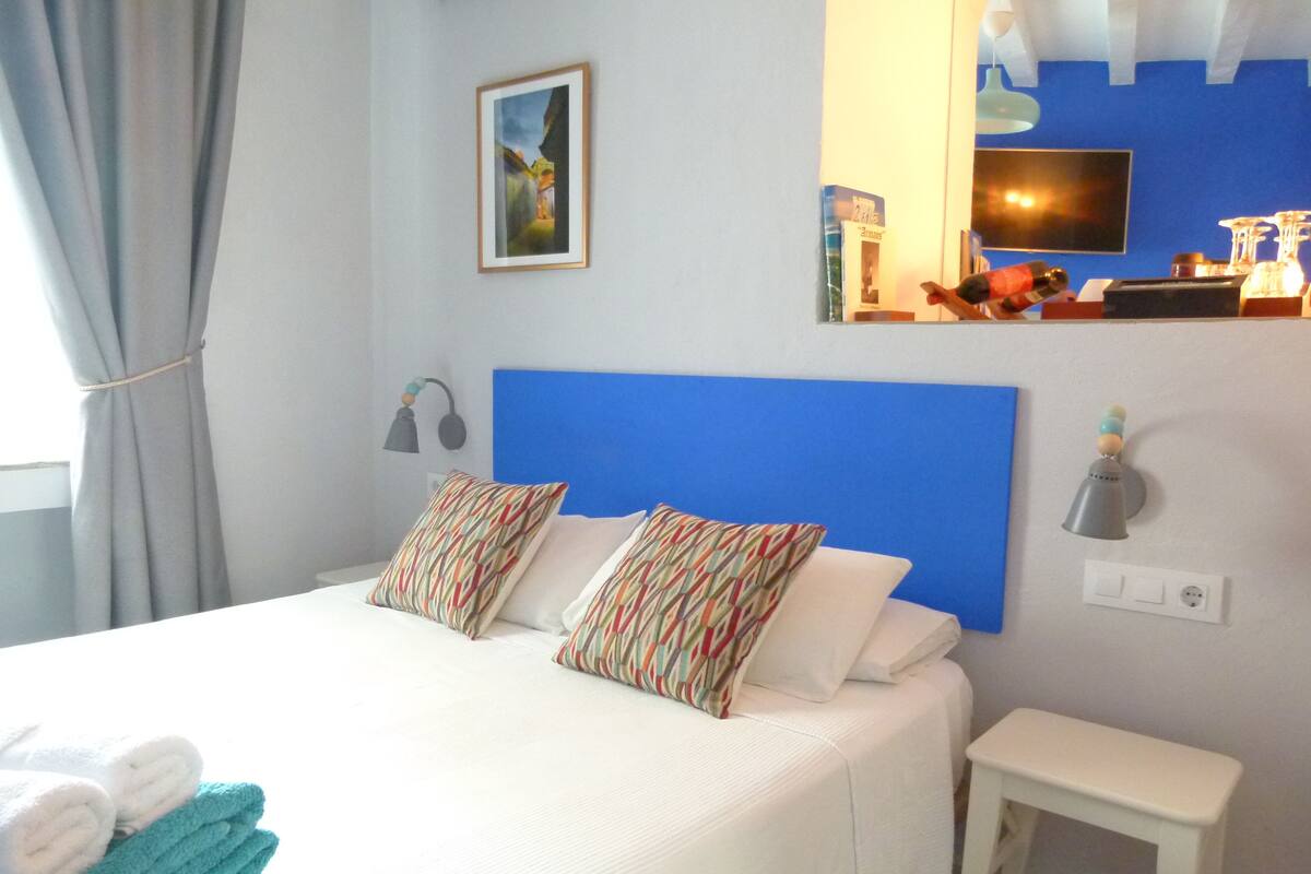 Ein ordentlich bezogenes Bett mit gemusterten Kissen, ein Nachttisch, eine kleine Nachttischlampe und ein an der Wand befestigtes Fernsehgerät.