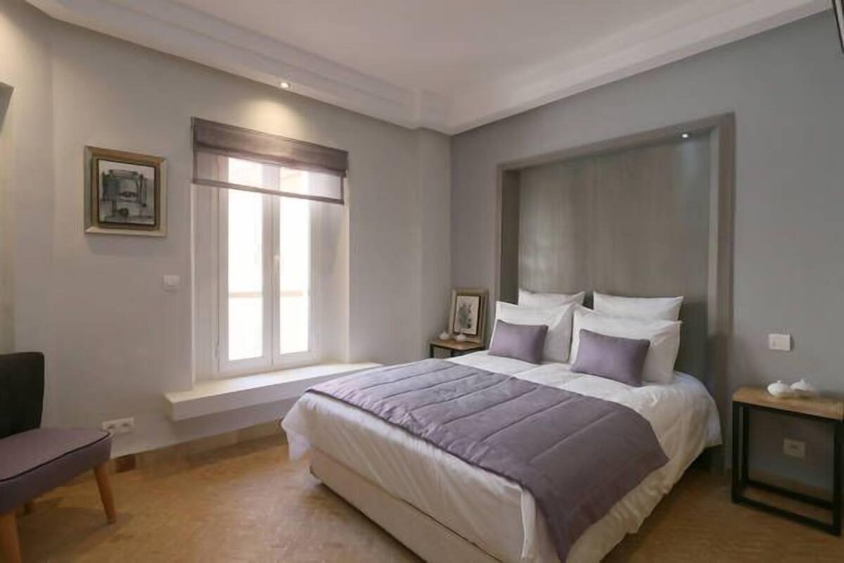 A bedroom with a large bed, a window with blinds, a bedside table, and a chair.