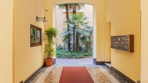 A hallway with a red carpet leading to a courtyard with palm trees and a view of a building. - Milan Retreats - Porta Venezia (Milan)
