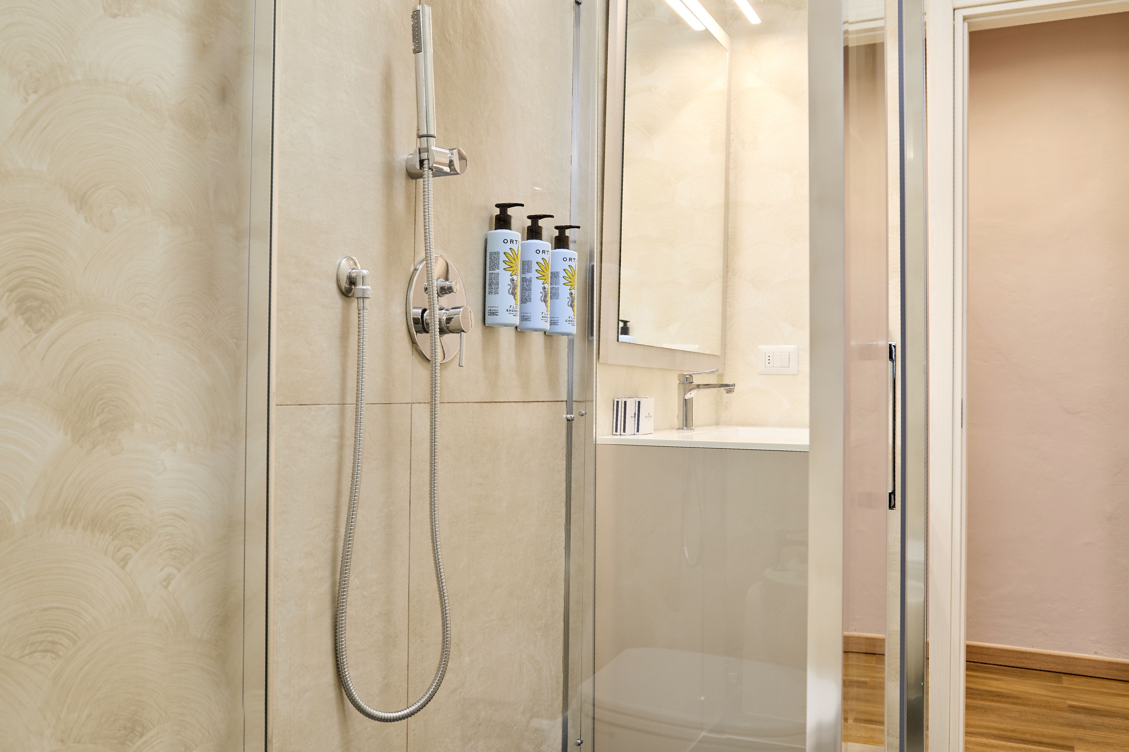 A modern bathroom with a glass shower enclosure, a handheld showerhead, and two bottles of body lotion on the wall.