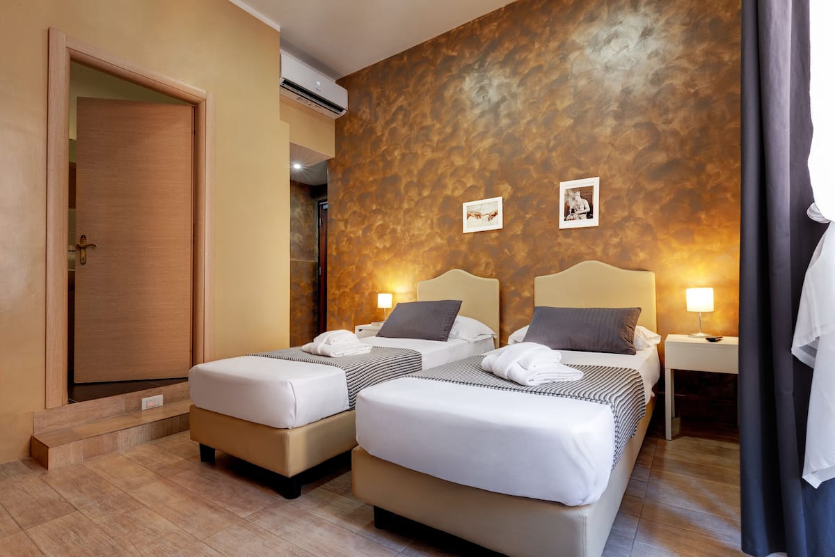 Habitación de hotel con dos camas, una puerta de madera, aire acondicionado montado en la pared y una pequeña mesa con lámpara.
