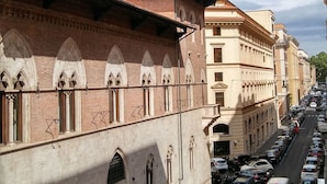 A street view with a row of parked cars and a building with arched windows and a tiled roof. - AmoRoma (Rome)