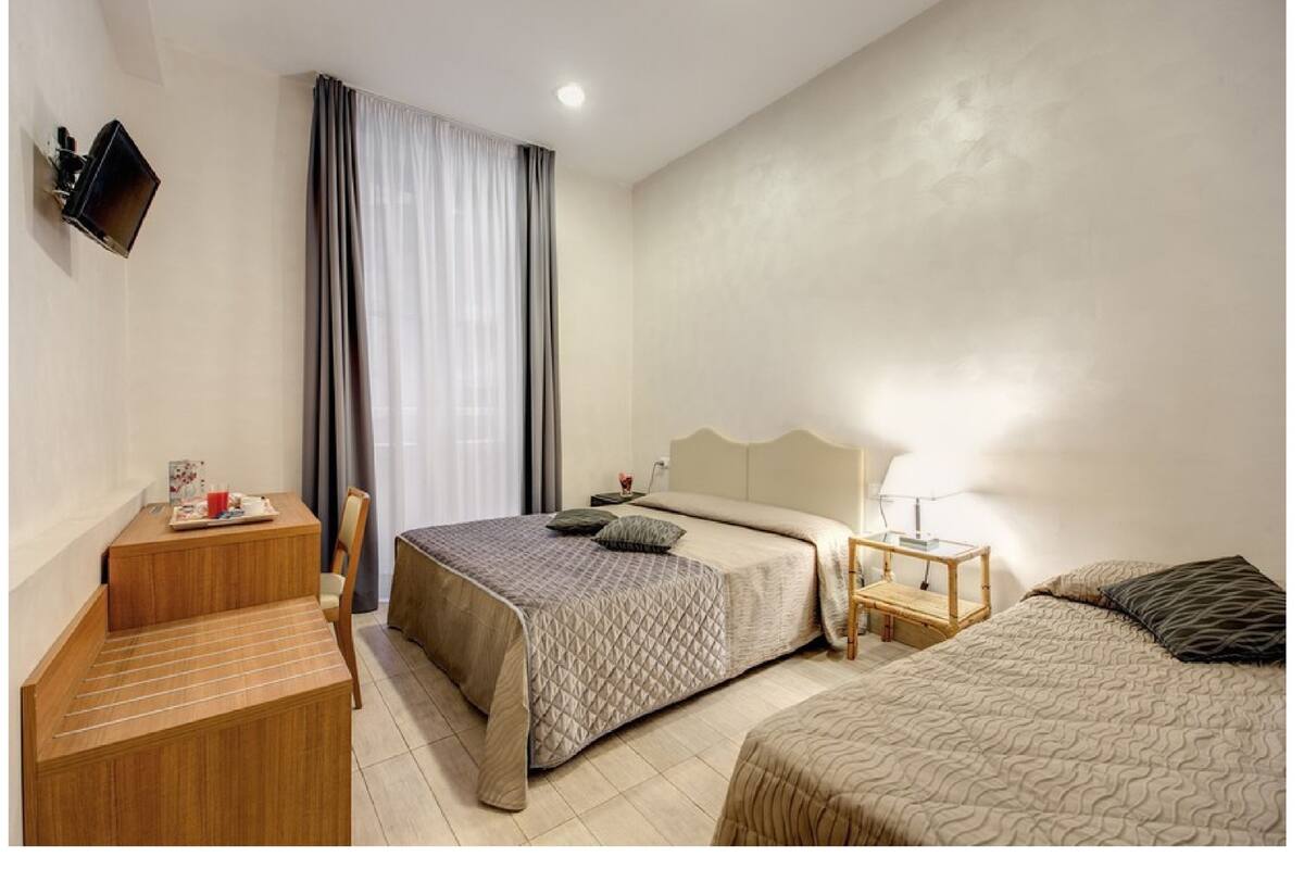 Habitación de hotel con cama, mesita de noche, lámpara, televisor montado en la pared y ventana con cortinas.